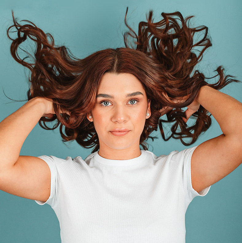 A woman with voluminous, wavy brown hair wearing a white ribbed t-shirt, holding her hair outwards against a solid teal-blue background.